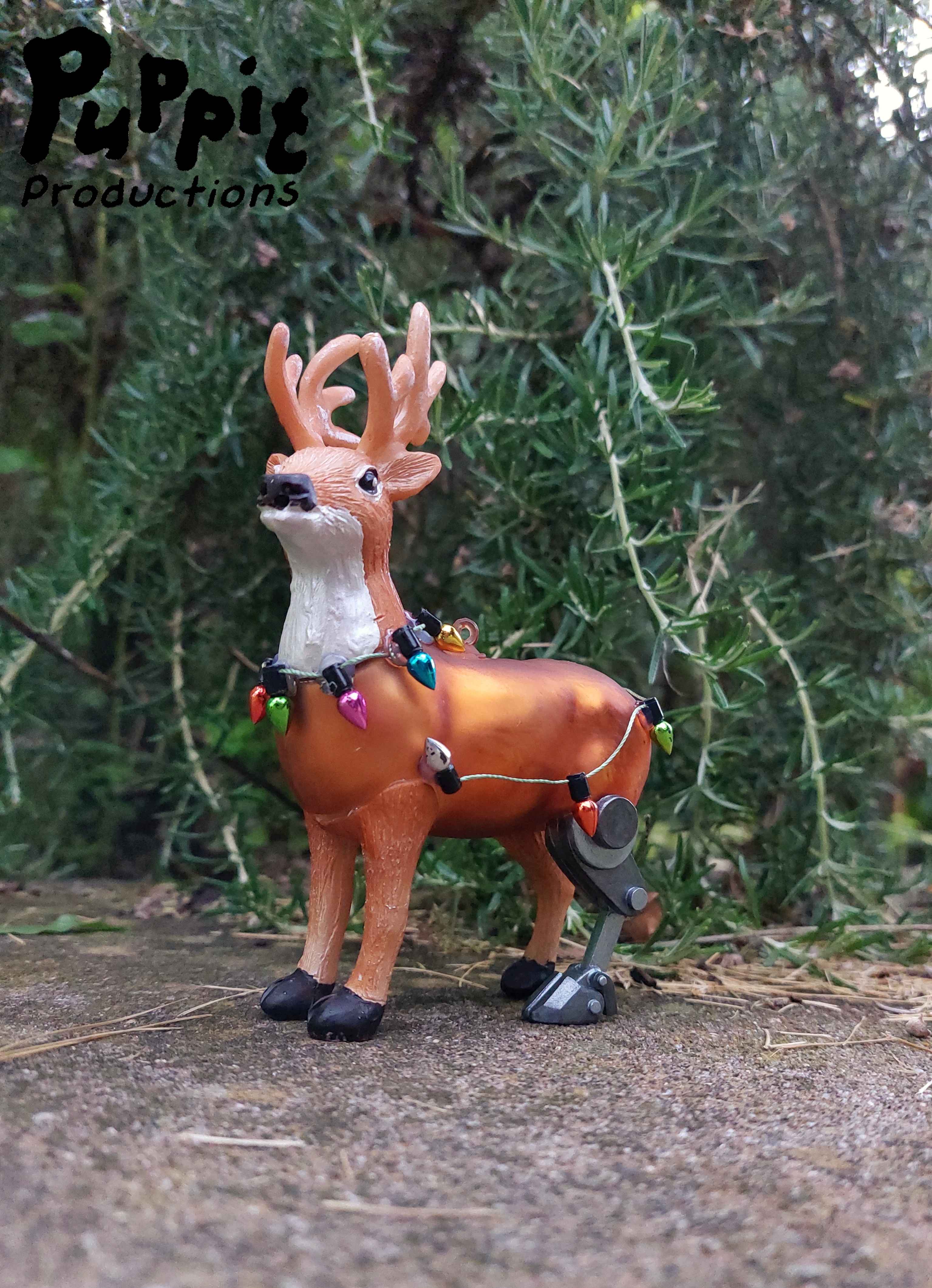 A picture of a reindeer christmas ornament with the left hind leg being a robotic looking prosthetic. The reindeer is standing outside in front of some greenery, and has miniature christmas lights wrapped around its torso.