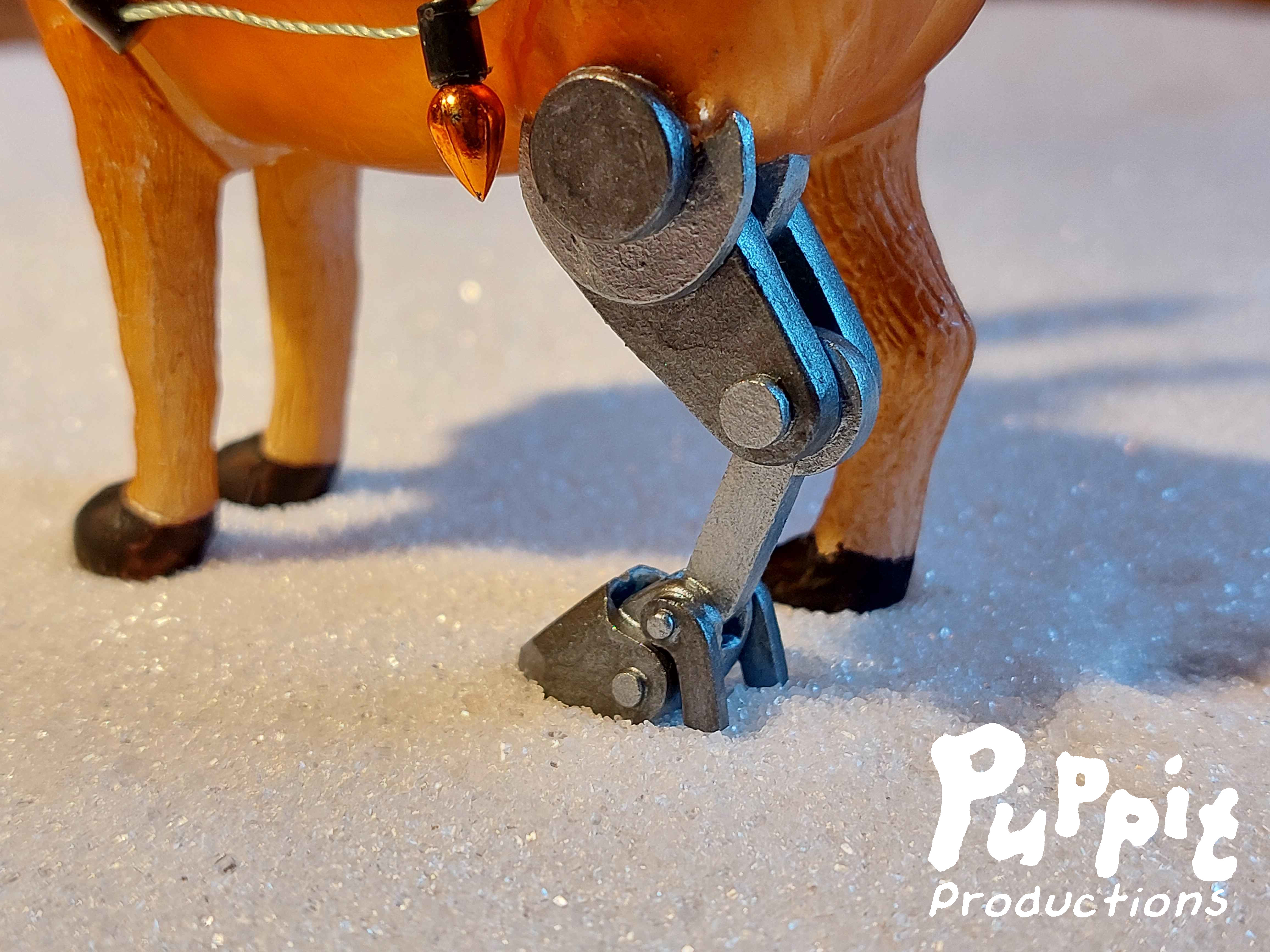 Close-up of the bottom half of a reindeer christmas ornament standing in snow. The left hind leg is a robotic looking prosthetic in silver metal tones.